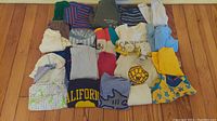 Overview of 25 pieces of toddler boys clothing laid out on floor including shirts, pants, shorts, and sweaters in assorted colors and patterns.