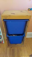 Front view of wooden bin holder with two blue plastic bins inside. Bin contents not included.