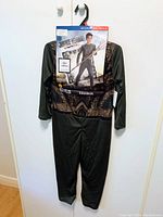 Aquaman costume hanging on wall with label featuring character image and packaging details, costume is dark green with gold printed armor details, long sleeved jumpsuit style.