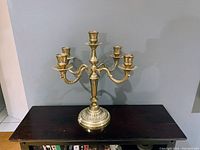 Brass five-arm candelabra shown standing on a dark wooden surface against a plain wall background. The candelabra has one central candle holder and four extending curved arms.