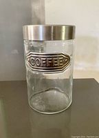 Clear cylindrical glass coffee container with metal lid and embossed 'COFFEE' metal plaque on front.