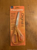 Photo shows Fiskars scissors in original orange packaging on wooden background. Packaging highlights features like sharper blades, perfect for fabric, ergonomic comfort, and 8-inch size.