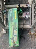 Image of the gas weed wacker with trimmer head, handle, debris guard, plastic bag with parts, and original green box on wooden steps.