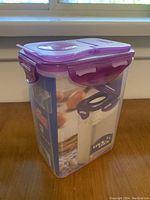 Front side view showing the transparent plastic container with a purple lid and locking clips, labeled with storage info and food images.