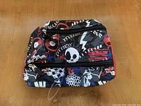 Large pencil case with multiple zippered compartments and black, white, red, and blue graffiti-style skull and lightning bolt print on a wooden surface.