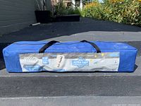 Blue carry case of the pop-up canopy with label showing features and brand, placed on pavement outdoors.
