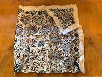 Silk scarf spread out on wooden surface showing detailed brown and black floral and scroll pattern on beige background