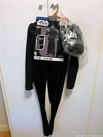 Black Kylo Ren child costume jumpsuit hanging with attached packaging and accompanying Kylo Ren mask in clear plastic packaging hanging beside it on a white door.