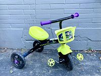 Side angle of toddler tricycle showing green seat, yellow Toy Story themed front basket, black frame, and three wheels on asphalt surface against gray brick wall.