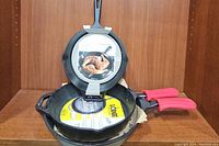 Three black cast iron skillets stacked in front of wooden shelf, showing the red silicone handle covers and original packaging.