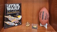 All lot items shown together on wooden shelf: Star Wars Episode I book, two small ship figurines (one clear acrylic cruise ship and one in a display case), and autographed Wilson NFL football on stand.