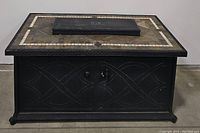 Front view of propane fire pit showing stone tile surface and black metal base with decorative patterns.