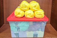 Plastic storage bin with a red lid containing assorted yarn skeins, topped with five yellow yarn skeins.