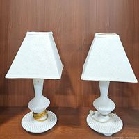 Pair of white vintage hobnail milk glass table lamps with white fabric shades sitting on a wooden surface against a wood background.