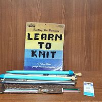 Learn to Knit booklet and assorted knitting needles arranged on wooden shelf.