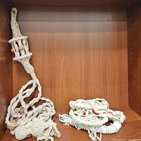 Two braided macrame plant hangers displayed folded inside a wooden shelf space. One hanger has natural wooden beads, the other has red beads.