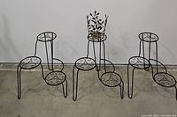 Three black metal 3-tier plant stands with decorative scrollwork on each tier. One metal floral planter holder with leaf motif seen on top of middle stand.