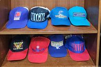 Front view of the first shelf of snapback hats including Phoenix Suns, Orlando Magic, Washington Wizards, and Charlotte Hornets hats on top row; and Clinton Store event, Washington Bullets, Baltimore Ravens, and Washington Bullets/Bucks hats on bottom row.