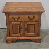 Front view of solid oak nightstand showing 1 drawer and 2 doors, wood grain and metal handles.