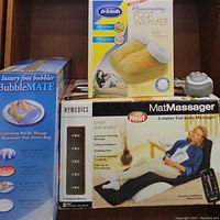 Photo of boxed Homedics MatMassager, Dr. Scholl's foot warmer slippers in box, Homedics Bubble Mate foot spa box, and Brookstone handheld massager device on shelf