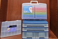 Photo showing four plastic storage containers arranged on wood surface: a clear bead box, blue multi-drawer box, rainbow storage carrier, and clear compartmentalized box