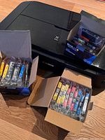 Canon Pixma printer with several open boxes showing XL ink cartridges in multiple colors, some still sealed in packaging, on wooden floor.