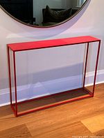 Full view of the red steel welded console table placed against a wall beneath a round mirror, showing its slim, rectangular open frame design and vibrant red color.