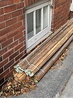 Bundles of finger joined framing lumber stacked outdoors beside a brick wall and a small basement window. Shows approximate length and condition with some discoloration.