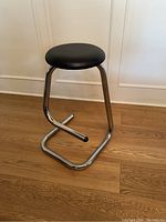 Side and front angle view of the black leather cushioned stool with chrome steel paperclip frame, showing the footrest and seat cushion.