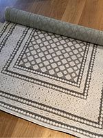 Rolled up outdoor woven rug on wooden floor showing the full geometric pattern in cream and gray colors.