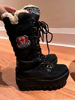 Side view of black Pajar tall boot with visible Sherpa lining, detailed side zipper, and front laces showing metal eyelets on a hardwood floor.