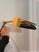 Picture light mounted on wall, showing cylindrical shade and brass finish with attached cord.