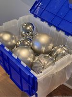 Top down view of a clear plastic bin containing 9 oversized silver Christmas balls with matte, glitter, and antique mirror finishes.