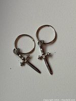 Pair of sterling silver hoop earrings each with a detailed sword charm pendant on a plain background.