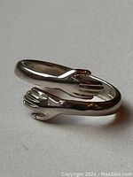 Close-up photo of the sterling silver ring shaped as two hands wrapping around finger, showing the ring band and hand details.