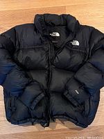 Front of black puffer jacket laid flat showing chest logos, sleeve “700” embroidery, zippered hand pockets