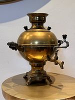 Front-left angle view of brass samovar kettle on a wooden surface showing spout, handle, and base details with a white wall background.