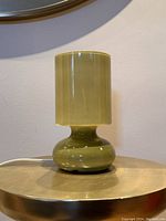Front view of the olive green glass lamp showing the cylindrical shade and rounded base on a metallic surface against a wall.