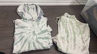 Folded hooded sweater and track pants laid flat on floor, showing bleach tie-dye green and white pattern