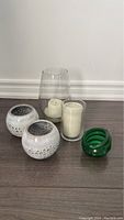 Photo of various candles and candle holders on wood floor, showing two white ceramic holders, one cylindrical glass holder with candle, two small white votive candles in glass, and a green glass holder.