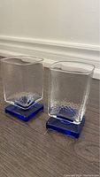 Photo of two small textured clear glass cocktail glasses with blue square bases on a wooden floor against a white baseboard wall.