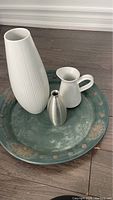 Photo showing large round decorative metal tray and three vases (tall white ceramic with ridges, small brushed metal, and small white pitcher-style ceramic)