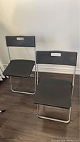 Two black IKEA folding chairs set up on a wood floor, showing front and seat view.