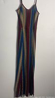 Full length sleeveless striped dress with spaghetti straps hanging against a white wall. The dress features vertical multicolor stripes with textured knit fabric.