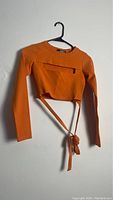 Photo of orange long sleeve cropped shirt hung on black hanger, showing both front and side views with wrap straps tied and untied.