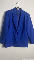 Front view of oversized bright blue blazer hanging on a hanger against a plain wall