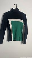 Black turtleneck sweater with central green panel bordered by white, hanging on wall hanger.