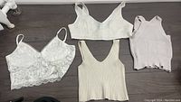 Four women's crop tops and bralettes in white, cream, beige, and light pink shades, featuring lace, knit, and ribbed textures, displayed flat