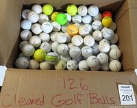 Box of 126 cleaned used golf balls showing many white balls with some yellow, green and orange golf balls spread out in the box.