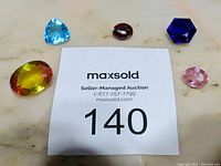 Five man-made faceted gemstones arranged on a marble surface around a MaxSold auction lot card with number 140.